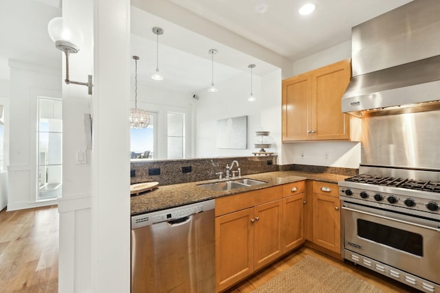 kitchen featuring wall chimney range hood, light wood-type flooring, dark stone countertops, stainless steel appliances, and a sink