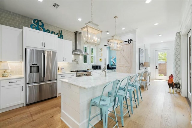 kitchen with a barn door, light wood-style flooring, wall chimney range hood, stainless steel fridge, and a center island with sink