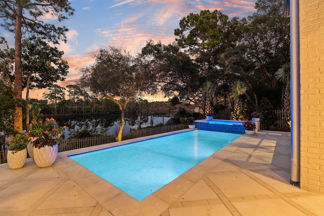 pool at dusk featuring a pool with connected hot tub, a water view, a patio, and fence