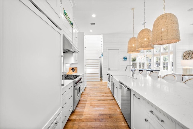 kitchen with stainless steel appliances, exhaust hood, white cabinets, light wood finished floors, and hanging light fixtures