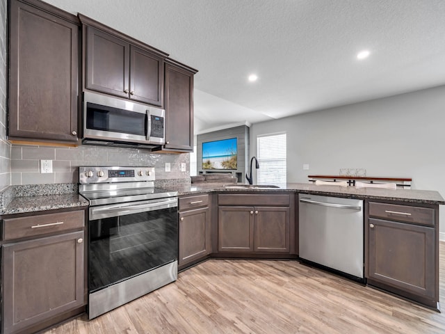 kitchen featuring light wood-style flooring, dark brown cabinetry, stainless steel appliances, a sink, and dark stone counters