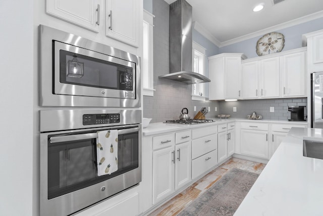 kitchen featuring stainless steel appliances, ornamental molding, light countertops, wall chimney range hood, and backsplash