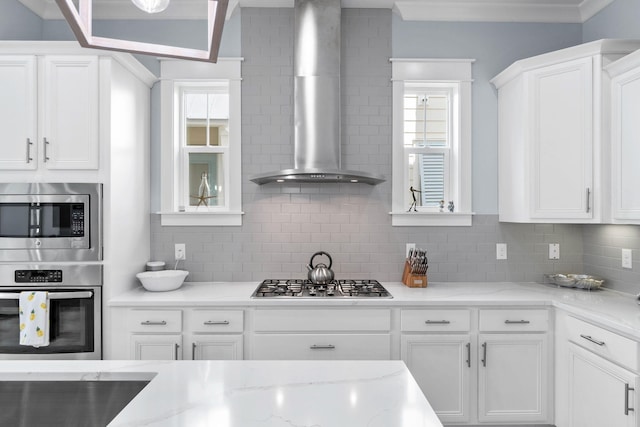 kitchen with ornamental molding, stainless steel appliances, white cabinets, wall chimney range hood, and backsplash