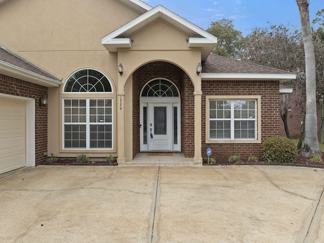 view of exterior entry featuring driveway, an attached garage, a shingled roof, stucco siding, and brick siding