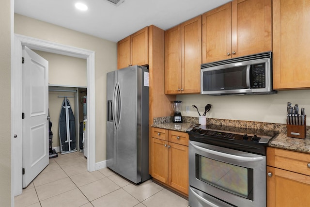 kitchen with brown cabinets, light tile patterned floors, dark stone counters, and stainless steel appliances