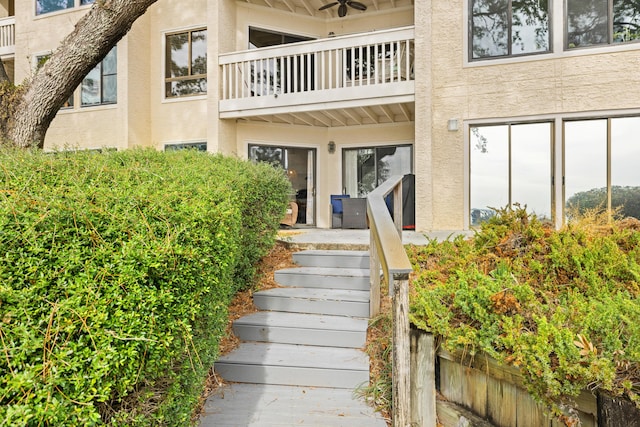 view of exterior entry with a ceiling fan, a balcony, and stucco siding