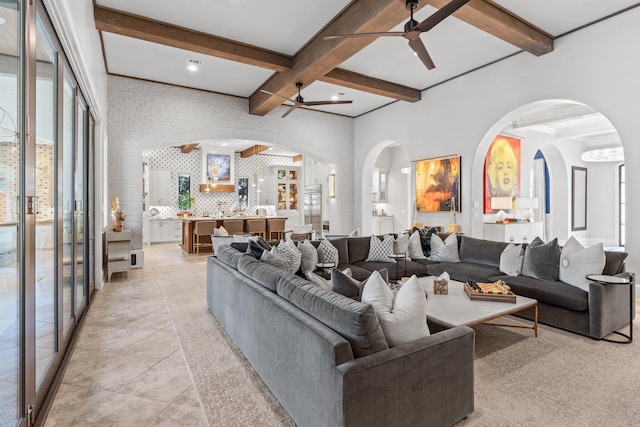 living area with arched walkways, light tile patterned floors, ceiling fan, brick wall, and beamed ceiling