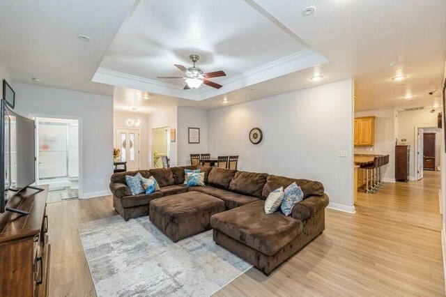 living area with baseboards, light wood-style flooring, ceiling fan, crown molding, and a raised ceiling