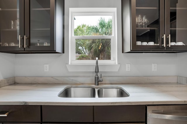 kitchen with glass insert cabinets, light stone countertops, dark brown cabinetry, stainless steel dishwasher, and a sink