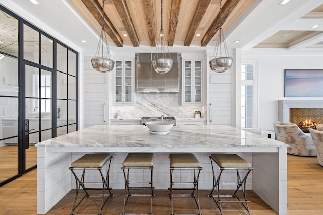 kitchen featuring beamed ceiling, light wood finished floors, backsplash, white cabinetry, and wall chimney range hood