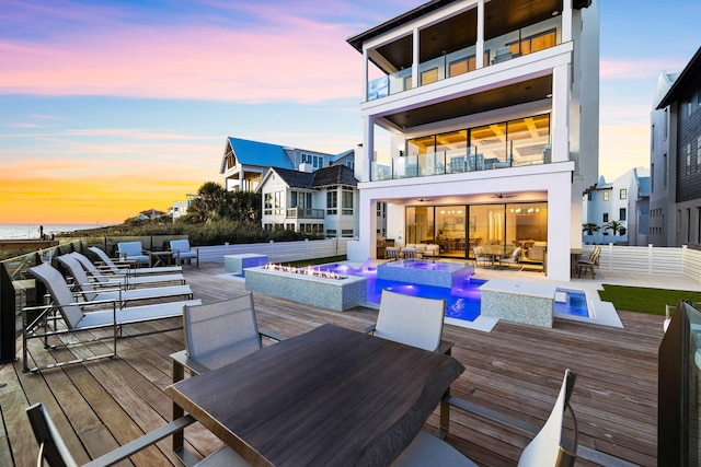 back of house at dusk featuring fence, outdoor dining area, a balcony, an outdoor pool, and an in ground hot tub