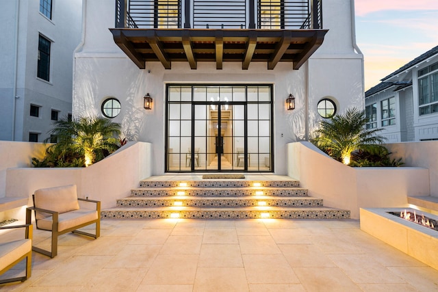 exterior entry at dusk with stucco siding, french doors, a balcony, and a patio