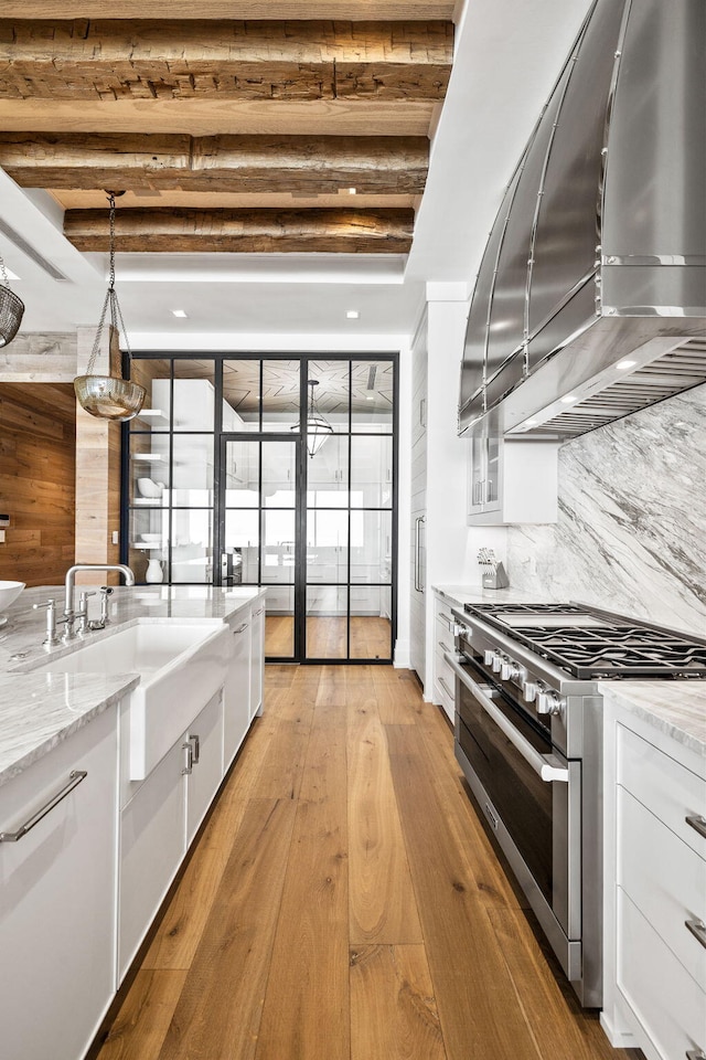 kitchen with beam ceiling, a sink, stainless steel stove, white cabinetry, and wall chimney exhaust hood