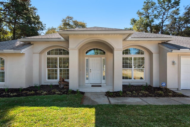 entrance to property with stucco siding, a lawn, and roof with shingles