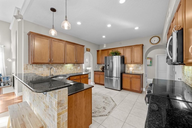 kitchen featuring brown cabinetry, dark stone counters, arched walkways, a sink, and appliances with stainless steel finishes