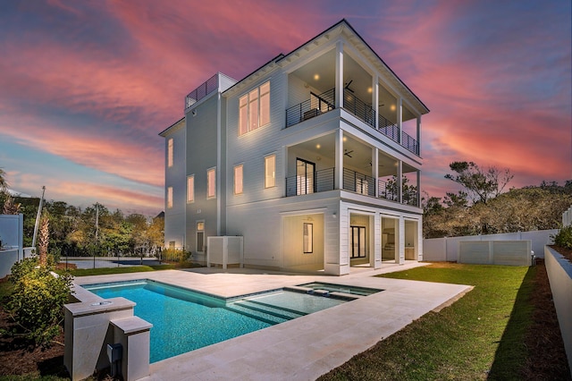 back of property at dusk with a balcony, a ceiling fan, a fenced backyard, a patio area, and a lawn