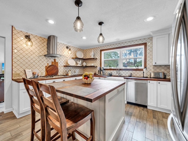 kitchen with a sink, appliances with stainless steel finishes, white cabinetry, wall chimney exhaust hood, and butcher block counters