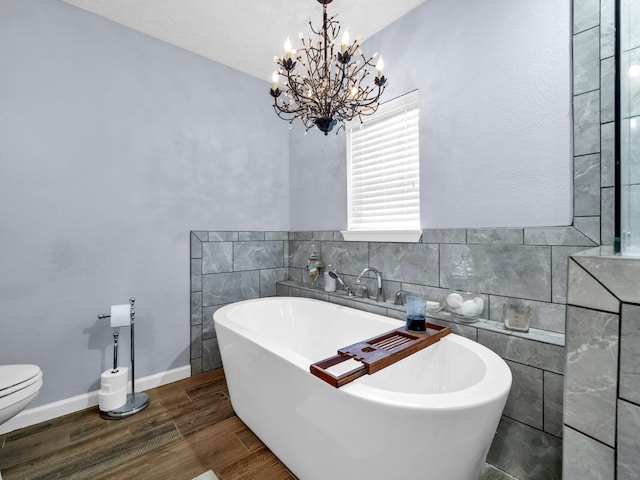 bathroom with toilet, wood finished floors, a freestanding tub, a notable chandelier, and tile walls