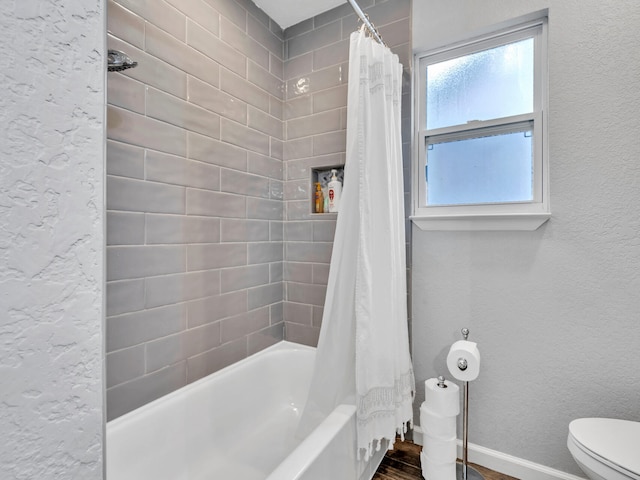 bathroom featuring toilet, a textured wall, shower / bath combo with shower curtain, and baseboards