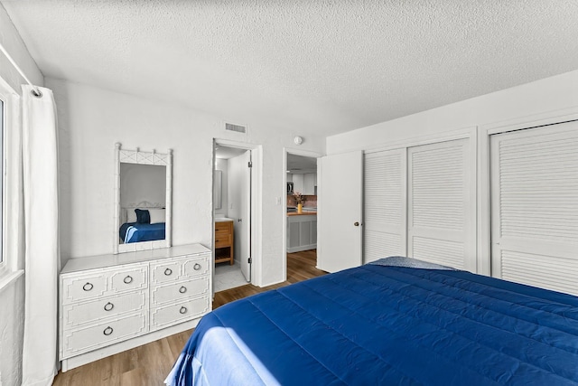 bedroom featuring wood finished floors, visible vents, ensuite bath, multiple closets, and a textured ceiling