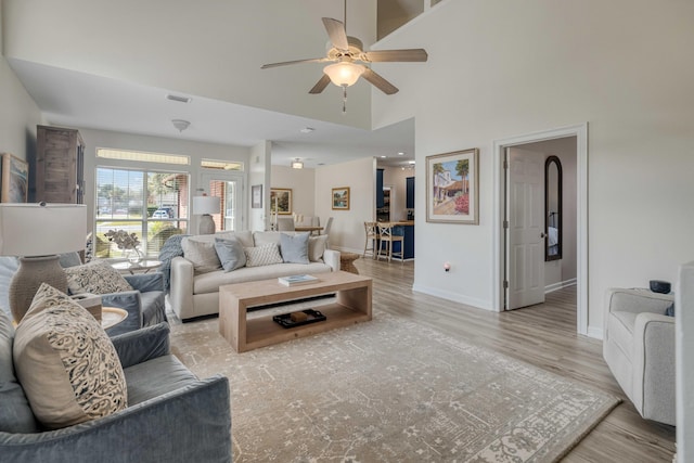 living area with a ceiling fan, visible vents, baseboards, light wood-style flooring, and a towering ceiling