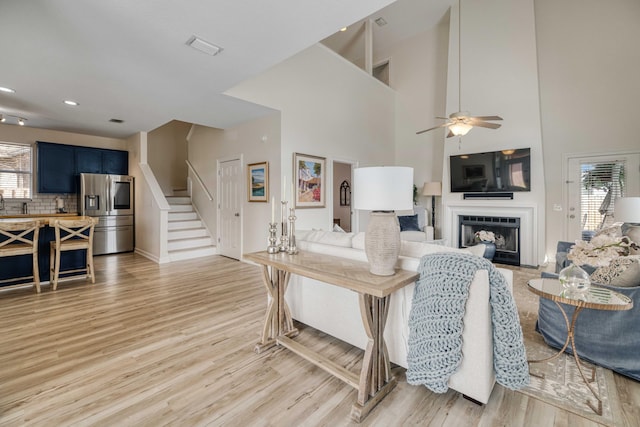 living area with light wood-style flooring, a high ceiling, a fireplace, ceiling fan, and stairs