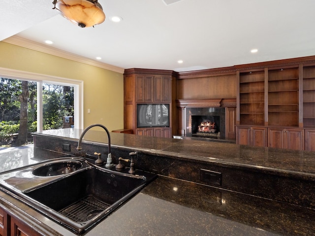 kitchen with crown molding, dark stone countertops, recessed lighting, a warm lit fireplace, and a sink