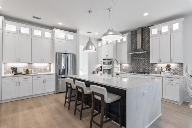 kitchen with appliances with stainless steel finishes, wall chimney exhaust hood, light wood-style floors, and a kitchen island with sink