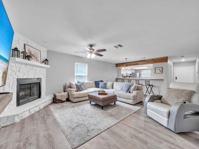 living area featuring a stone fireplace, ceiling fan with notable chandelier, light wood-style floors, and visible vents