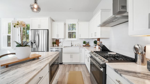 kitchen featuring a sink, light stone counters, stainless steel appliances, wall chimney range hood, and a healthy amount of sunlight