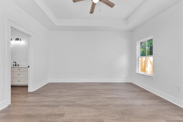 empty room with light wood-type flooring, a sink, baseboards, a raised ceiling, and ceiling fan