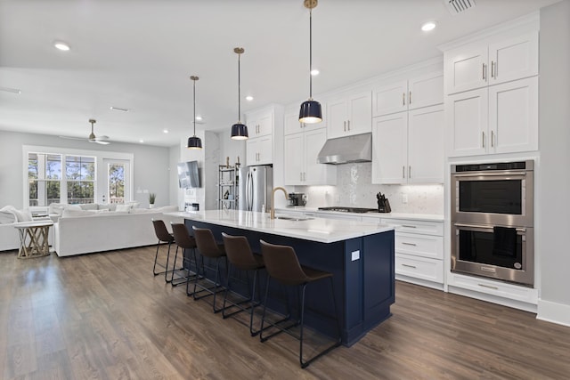 kitchen with a sink, under cabinet range hood, open floor plan, appliances with stainless steel finishes, and dark wood-style flooring