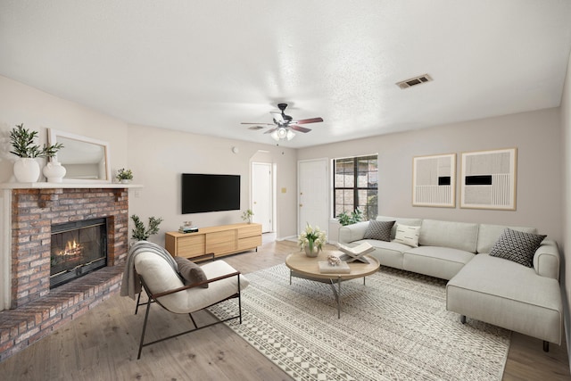 living area featuring a ceiling fan, wood finished floors, visible vents, a textured ceiling, and a brick fireplace