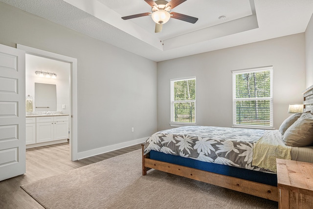 bedroom with a raised ceiling, a ceiling fan, ensuite bathroom, light wood finished floors, and baseboards