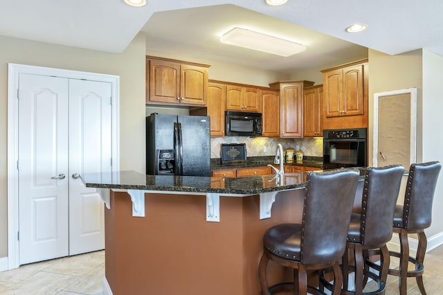 kitchen featuring tasteful backsplash, dark stone counters, an island with sink, a kitchen bar, and black appliances