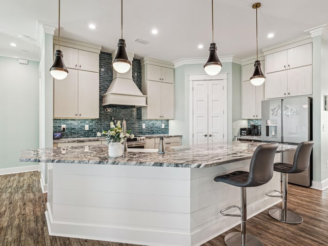 kitchen with a large island with sink, appliances with stainless steel finishes, dark wood finished floors, custom range hood, and decorative backsplash