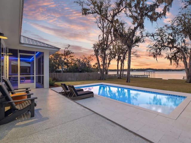 pool at dusk with fence, a patio, a water view, a fenced in pool, and a yard