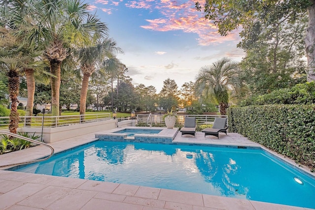 pool at dusk featuring a pool with connected hot tub and fence