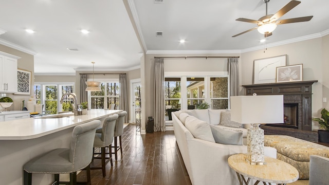 living area with a brick fireplace, visible vents, ceiling fan, ornamental molding, and dark wood-style flooring