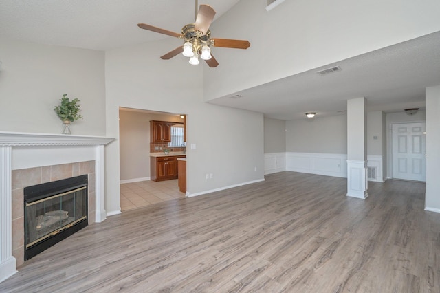 unfurnished living room featuring a wainscoted wall, a tiled fireplace, light wood finished floors, visible vents, and ceiling fan