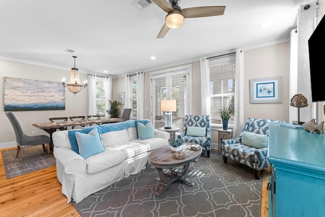 living room featuring crown molding, wood finished floors, baseboards, ceiling fan with notable chandelier, and visible vents