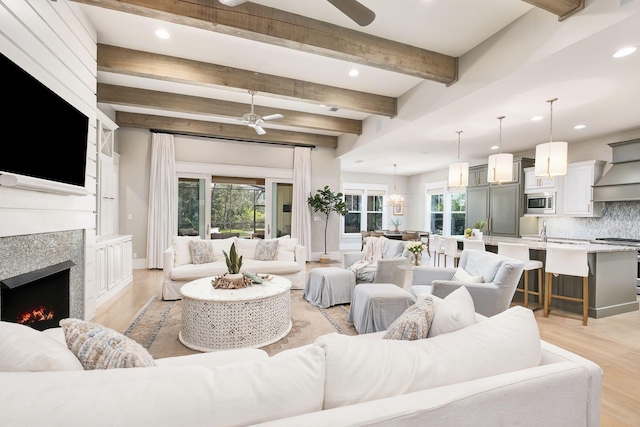 living area featuring beam ceiling, a lit fireplace, light wood finished floors, and ceiling fan