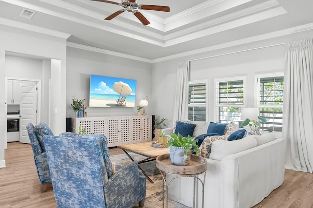 living room featuring crown molding, light wood-type flooring, a ceiling fan, visible vents, and a raised ceiling