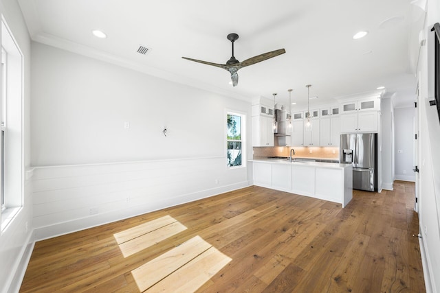 kitchen featuring ornamental molding, wall chimney range hood, a peninsula, stainless steel refrigerator with ice dispenser, and visible vents
