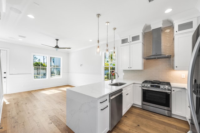 kitchen featuring a sink, wall chimney range hood, appliances with stainless steel finishes, a peninsula, and a wainscoted wall