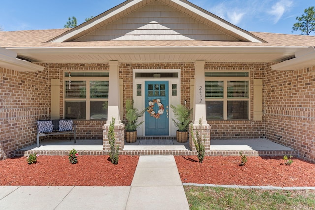 view of exterior entry featuring covered porch, a shingled roof, and brick siding