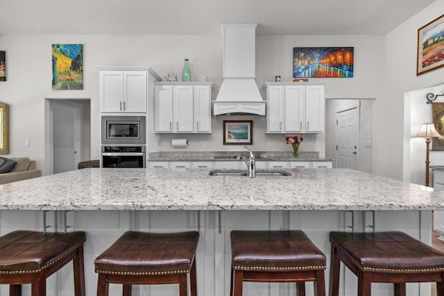 kitchen with white cabinets, a sink, custom range hood, light stone counters, and appliances with stainless steel finishes