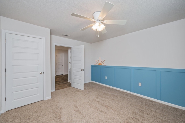 unfurnished bedroom featuring wainscoting, carpet floors, a textured ceiling, a ceiling fan, and visible vents