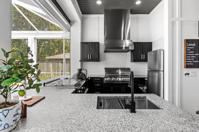 kitchen featuring freestanding refrigerator, light stone countertops, dark cabinets, tasteful backsplash, and wall chimney range hood