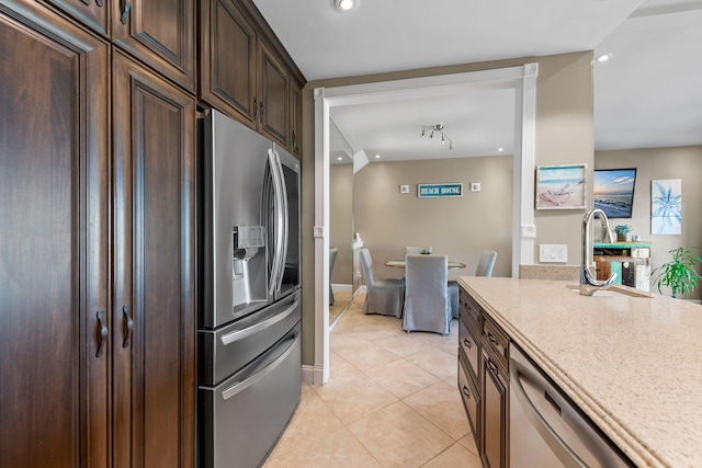 kitchen with appliances with stainless steel finishes, dark brown cabinets, a sink, light tile patterned floors, and light stone counters